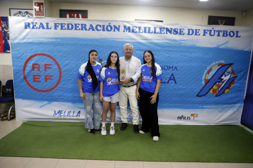 Four people pose in front of a blue Melillense football banner; three young women in blue shirts and a man in a white shirt hold a wooden award plaque/or trophy in the center.