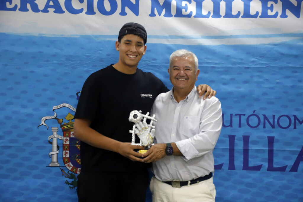 Young man in a black T-shirt and cap posing with an older man while holding a soccer-themed trophy in front of a blue banner.