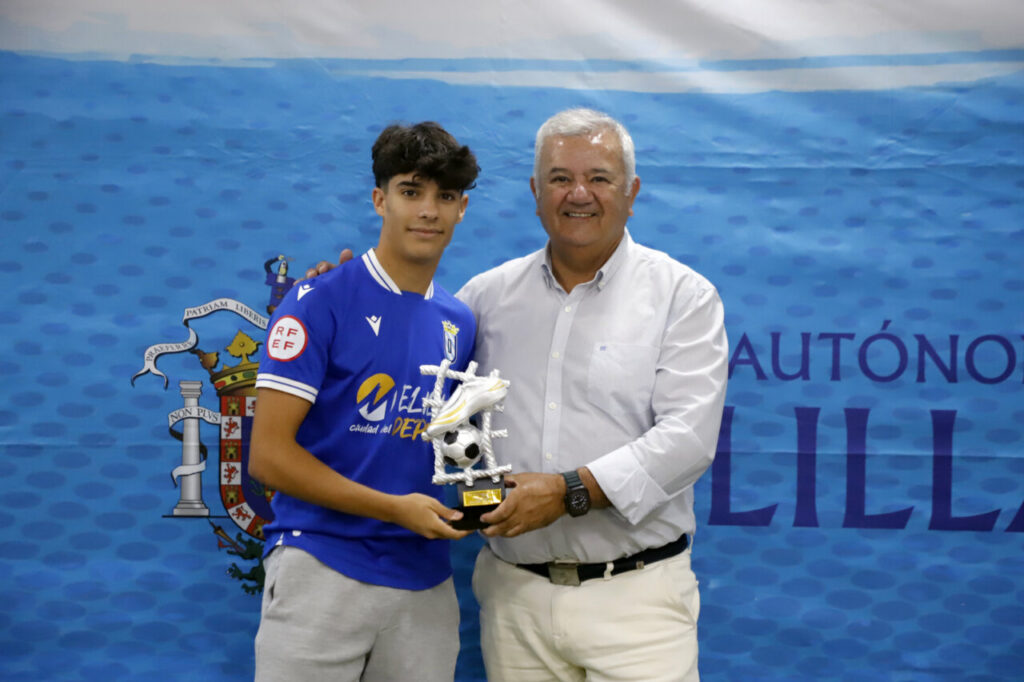 Young soccer player in a blue jersey poses with an older man, holding a trophy, in front of a blue event backdrop.