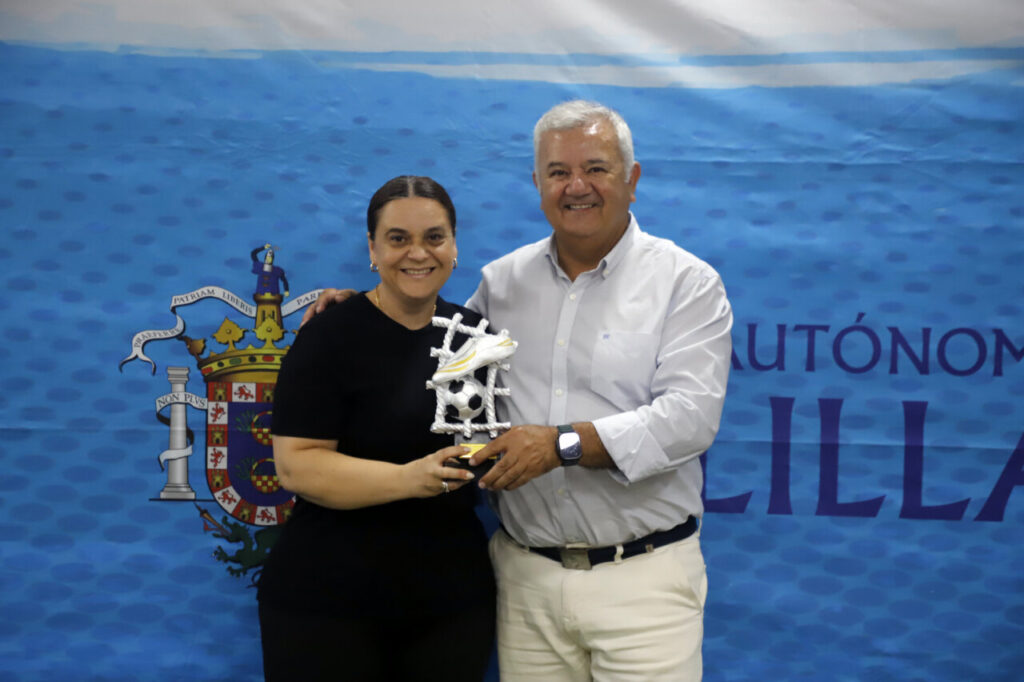 Man and woman posing with a soccer trophy in front of a blue backdrop featuring a crest and text.