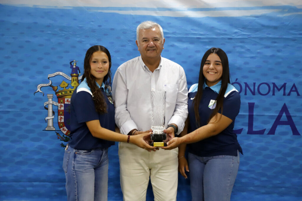 Man flanked by two young women presenting a crystal trophy at a blue event backdrop with a coat of arms.
