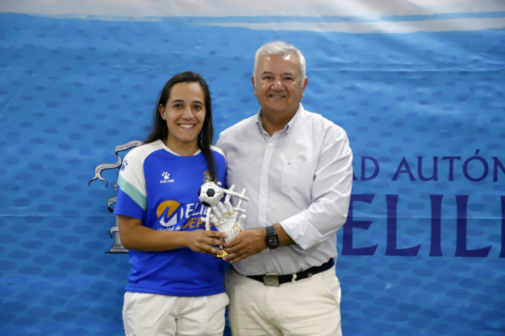 Smiling woman in a blue soccer jersey receives a trophy from a man in a light shirt at a sports award ceremony, with a blue patterned backdrop.
