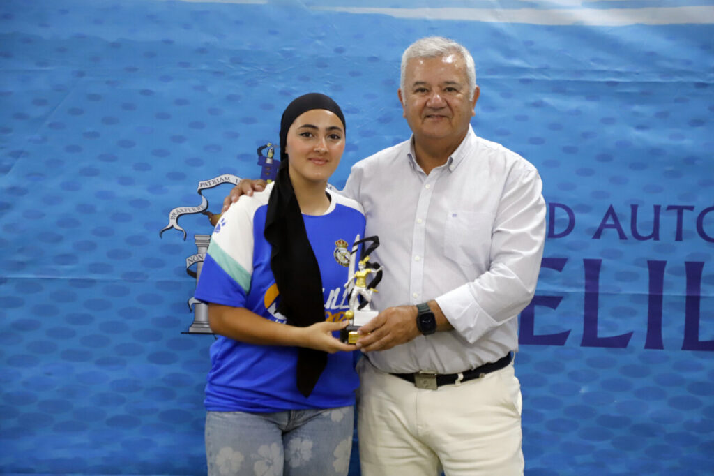 Young woman in a hijab and blue shirt holds a trophy, posing with a man at an awards backdrop.