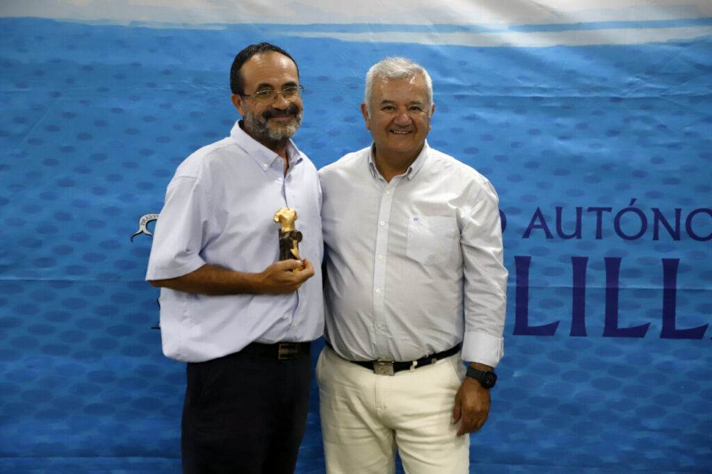 Two men posing at an awards event; the man on the left holds a small gold trophy.
