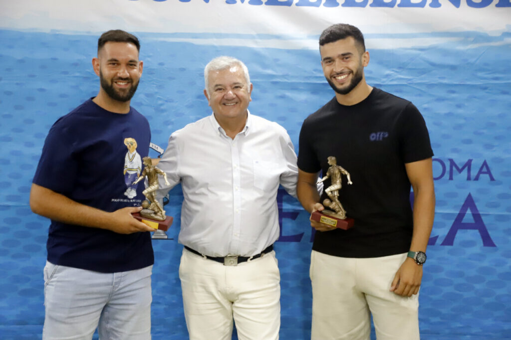 Three men pose together at an awards event, each holding a small bronze trophy shaped like a soccer player, in front of a blue backdrop.