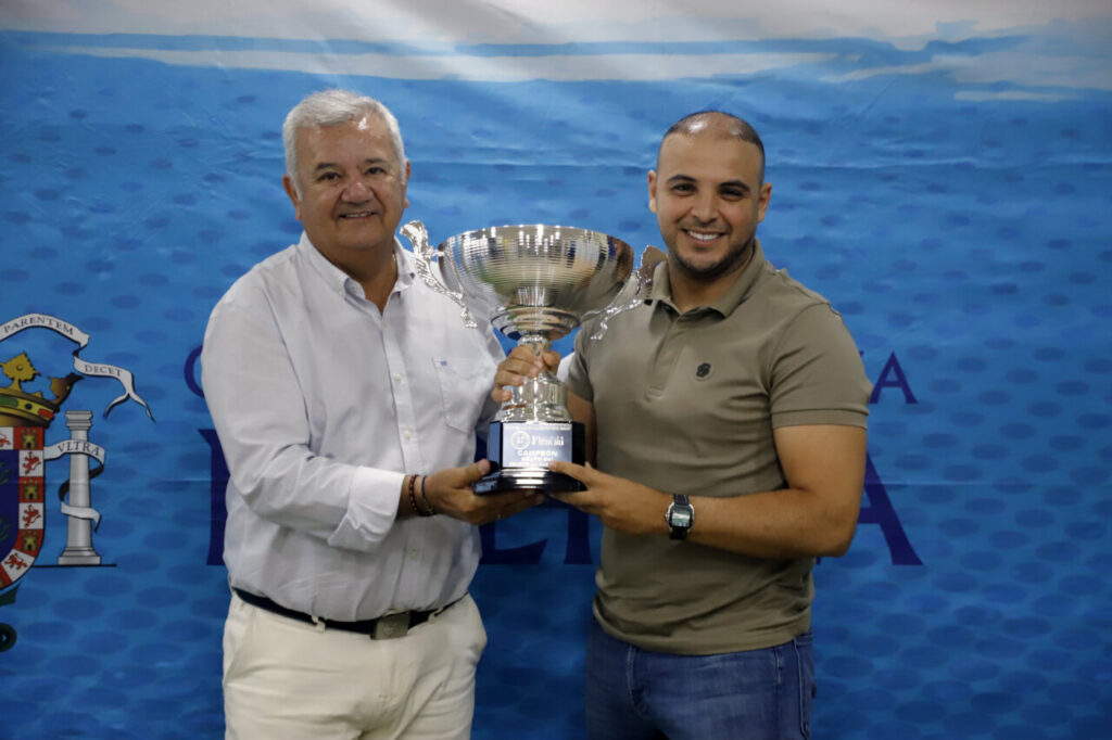 Two men pose together holding a large silver trophy with a blue base during an award ceremony, against a blue backdrop with a crest on the left.