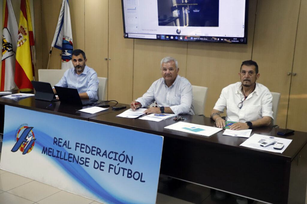 Three men sit at a conference table with laptops and papers, in front of a banner that reads Real Federación Melillense de Fútbol.