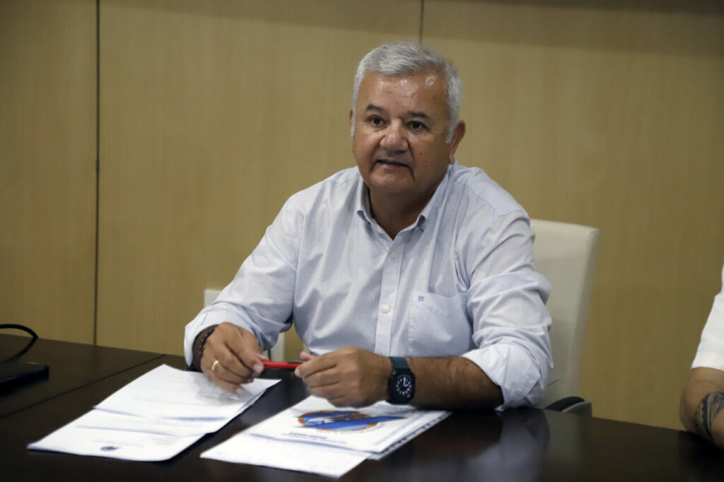 Man in a light blue button-down shirt sits at a dark conference table, holding a red pen over documents during a meeting in a the office.