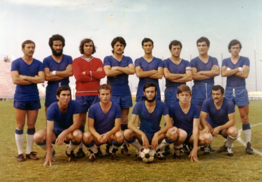 Vintage team photo of twelve men in blue soccer kits on a field, one in red goalkeeper jersey, posing with a ball at their feet.