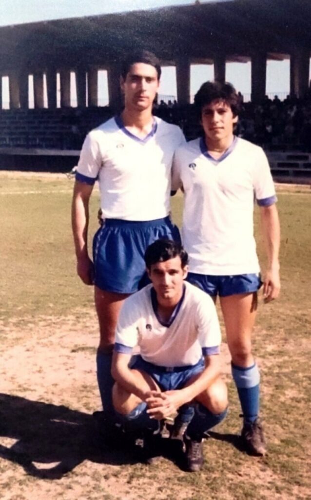 Three male soccer players in white and blue kits pose on a dusty field, two standing behind one crouching, with bleachers in the background.