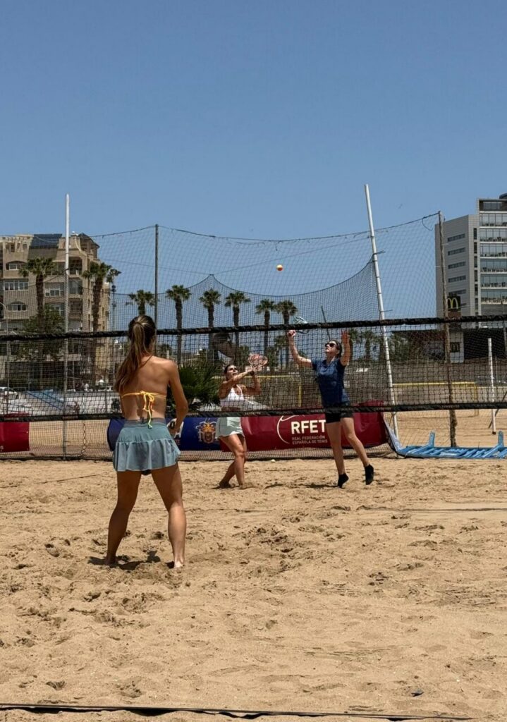 Three people play beach volleyball on a sunny sandy court with a net and urban backdrop; one player jumps to hit the ball over the net while others watch.