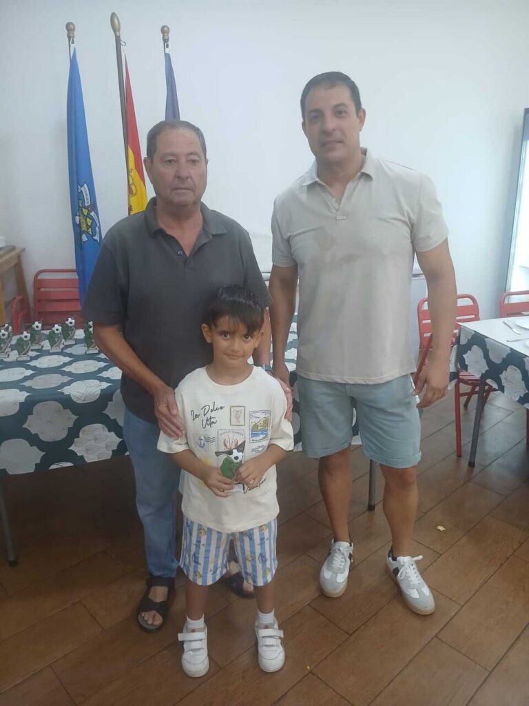 Three people posing indoors: a child in front, two men behind, with flags and tables in a meeting room.