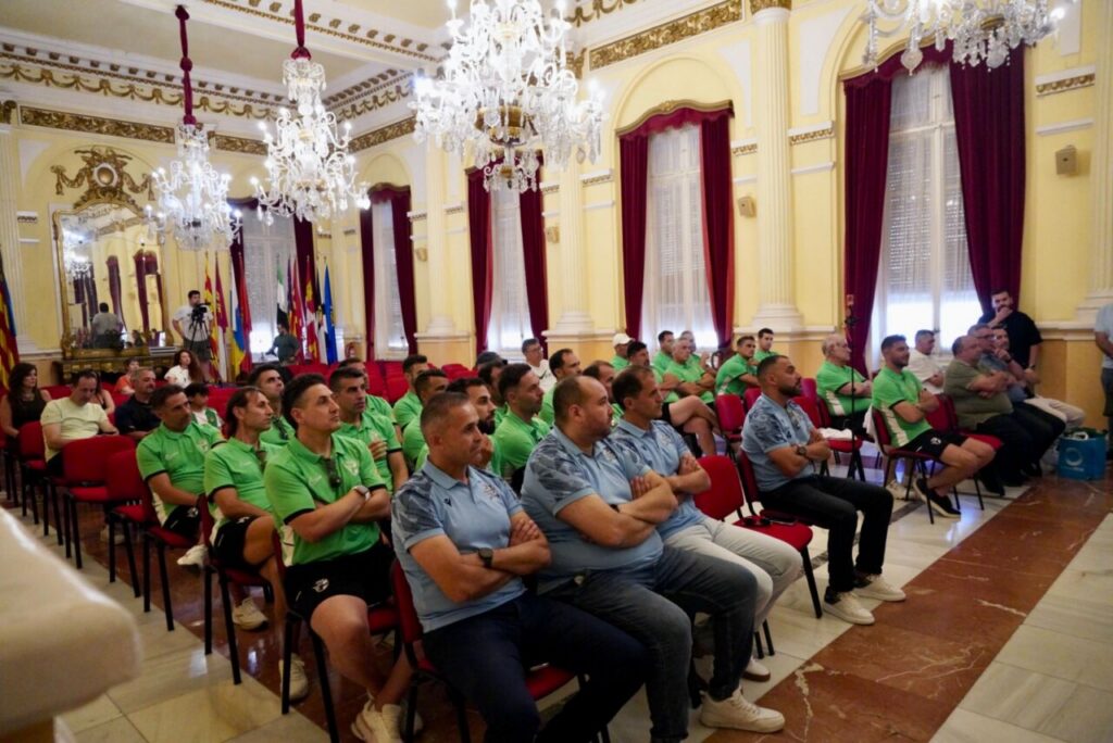 Group of men in green and blue polo shirts seated in red chairs in an ornate hall with chandeliers, attending a formal meeting or presentation.
