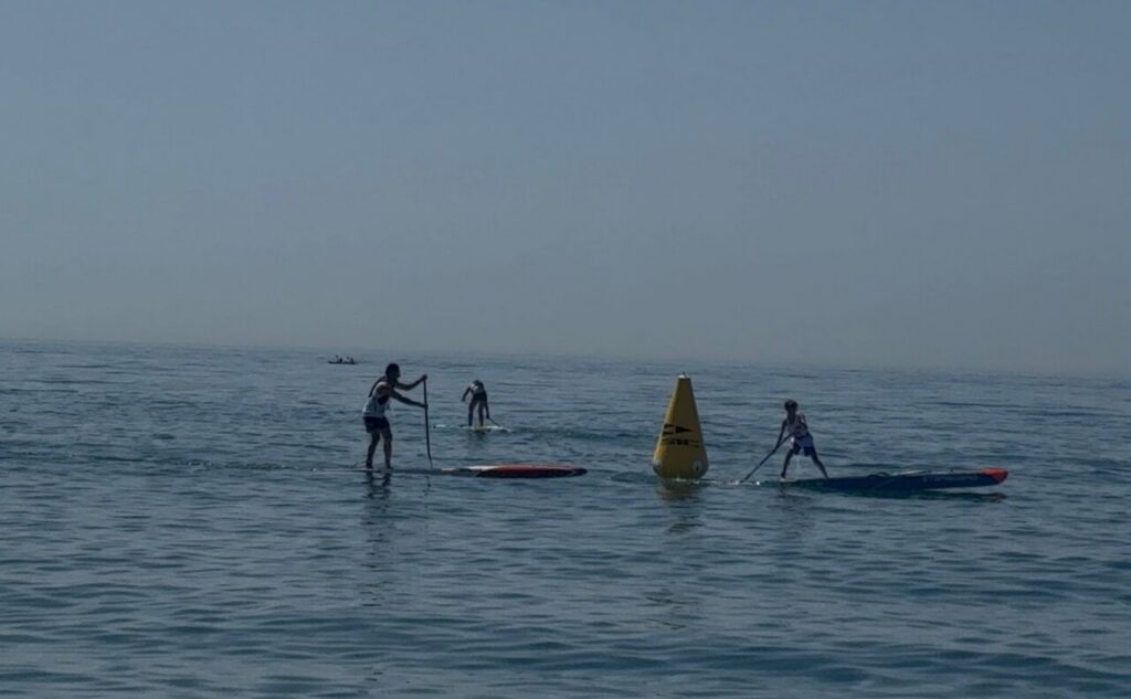 Three people stand-up paddleboarding on calm ocean near a yellow buoy at center distance edge of view.