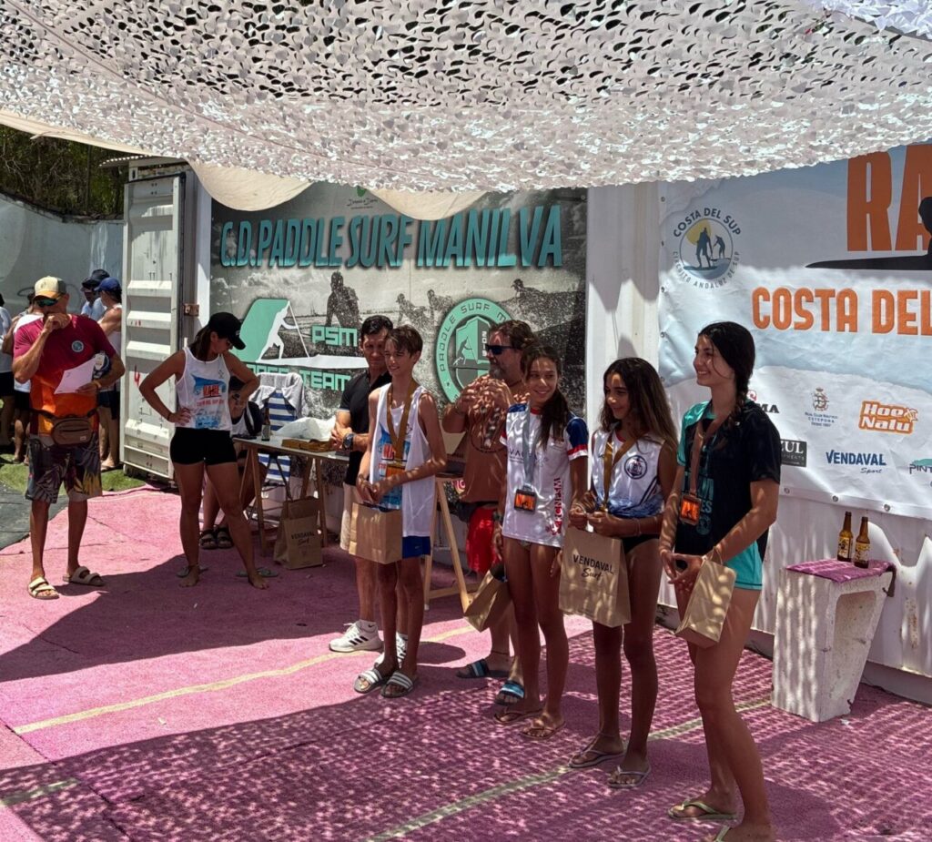 Group of young surfers with medals posing for a photo at an awards area under a camouflage canopy, banners and sponsors in the background.