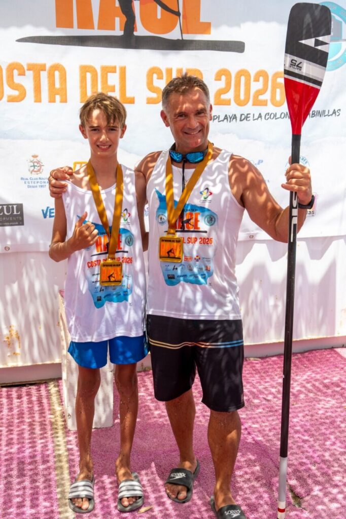 Man and boy with medals posing after a paddle racing event, holding a paddle together in front of a banner and pink ground cover.