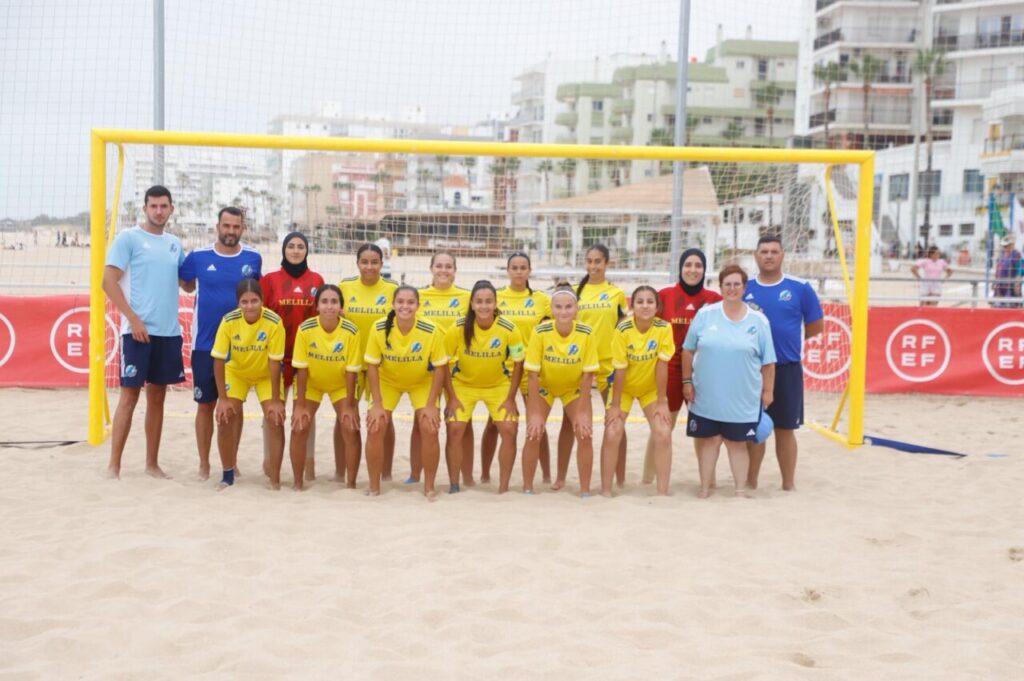 Women’s beach soccer team in yellow uniforms kneeling in front of a goal on the sand, with coaches in blue behind them.
