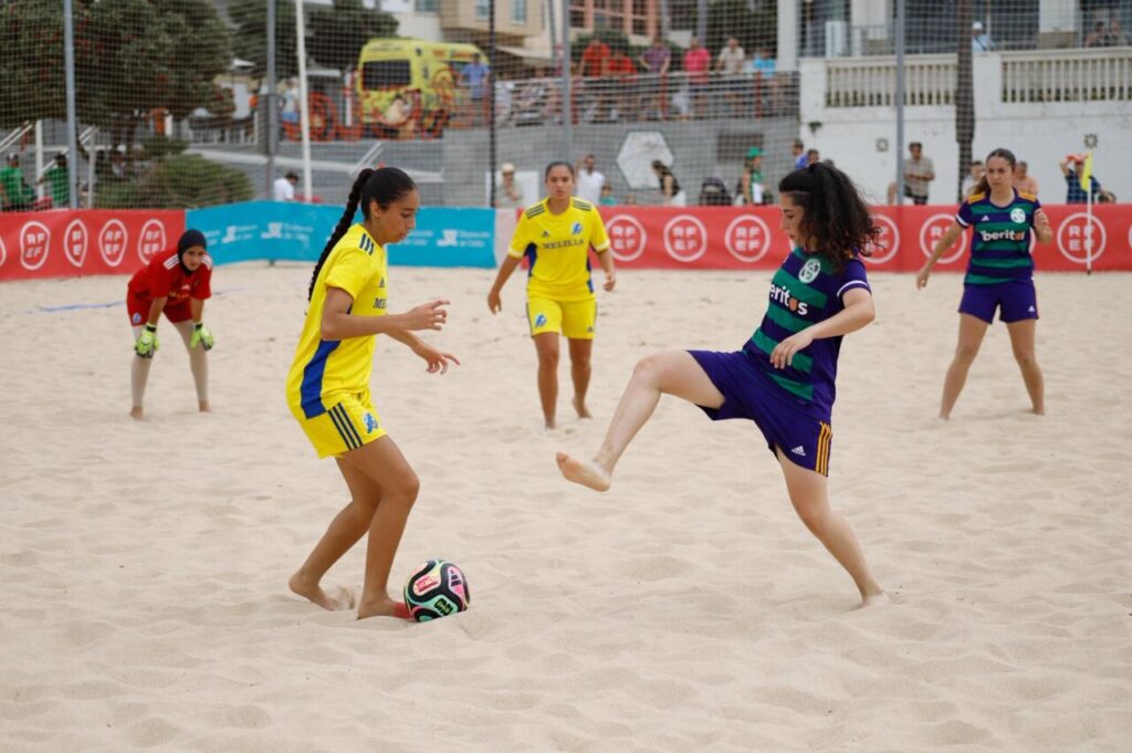 Women playing beach soccer on a sandy field; player in yellow controls the ball while a purple-team opponent kicks toward her.