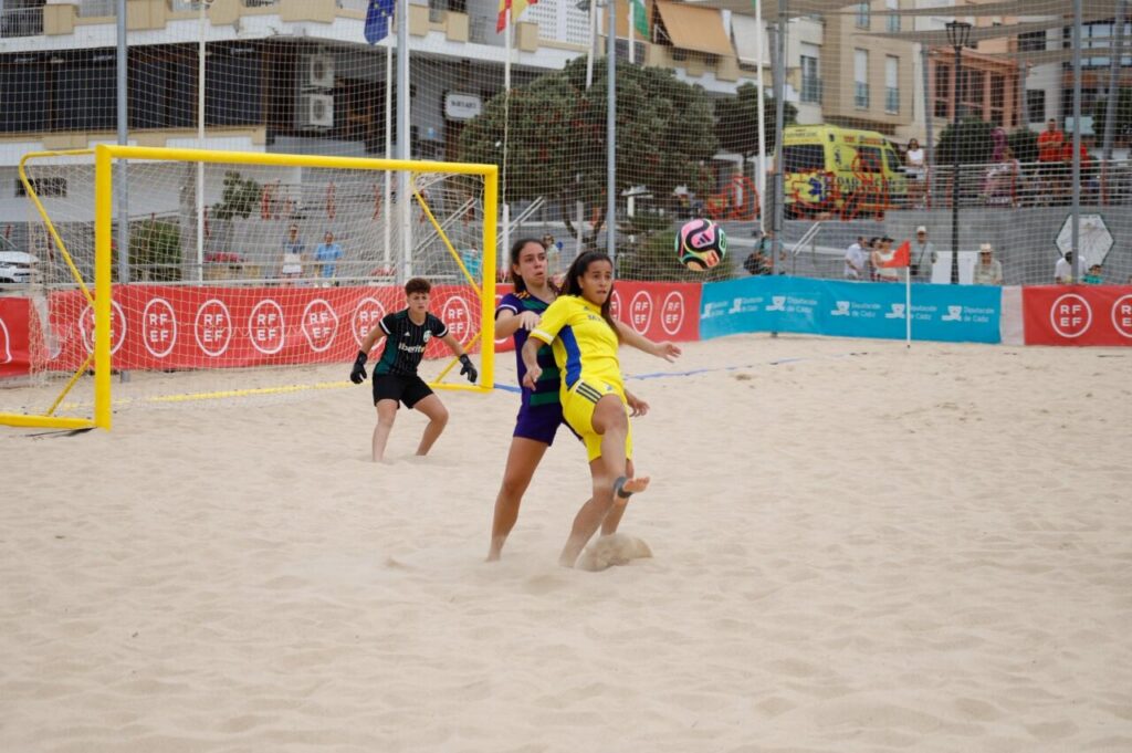 Youth beach soccer scene: a yellow-clad player kicks the ball near the goal as a purple-clad defender and goalkeeper react on sandy pitch.
