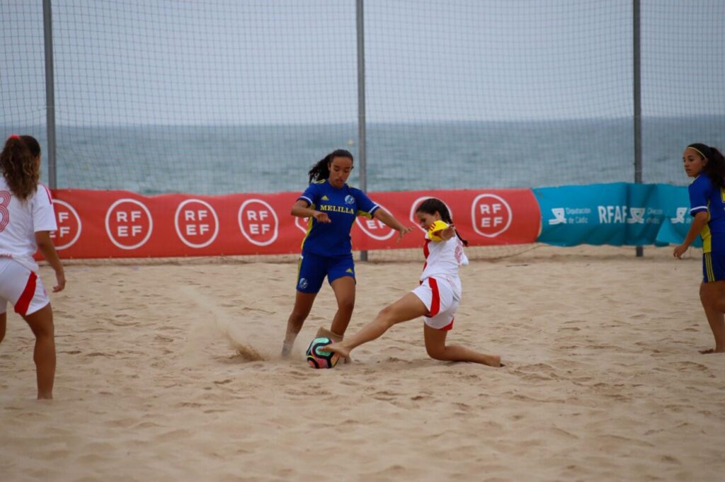 Girls' beach soccer match: one player in blue (Melilla) challenges a white-team player for the ball on sandy field near the ocean fence.