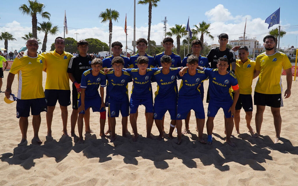 Group photo of a beach soccer team on sand, blue uniforms with 'MELILLA', flanked by coaches in yellow, under palm trees and flags.