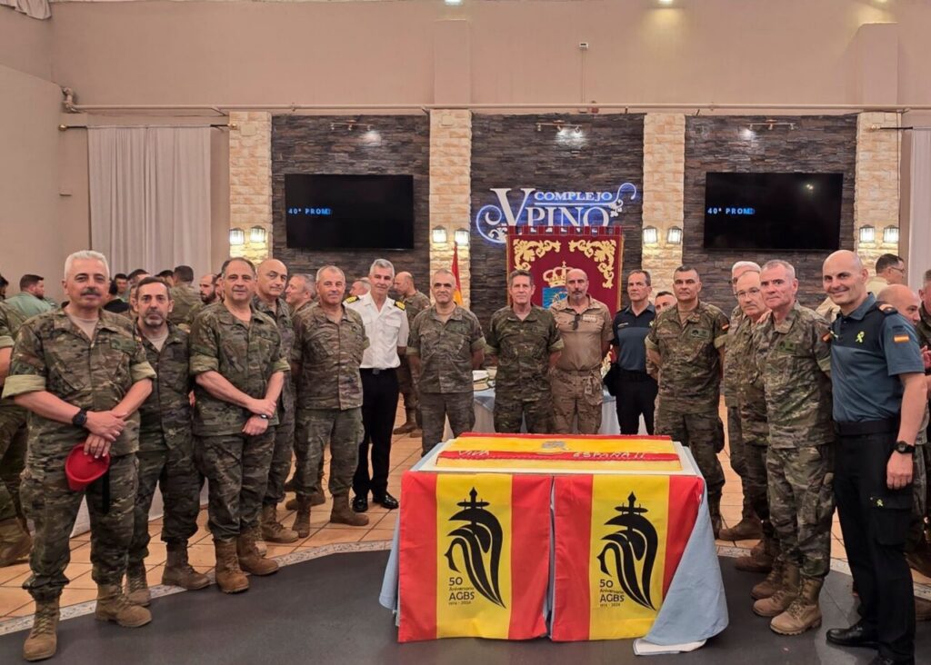Group of soldiers in uniform posing around a large celebratory cake with red-yellow panels and a black emblem, in a dining hall.