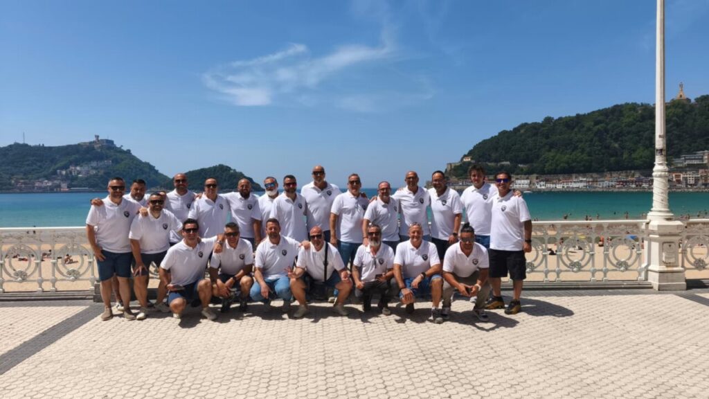 Group of men in white polo shirts posing on a sunny seaside promenade with turquoise water and green hills in the background.