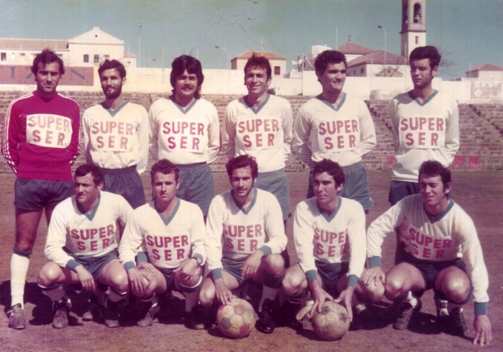 Vintage soccer team posing on a dirt field; white jerseys say 'SUPER SER', eleven players arranged in two rows with a stadium backdrop.