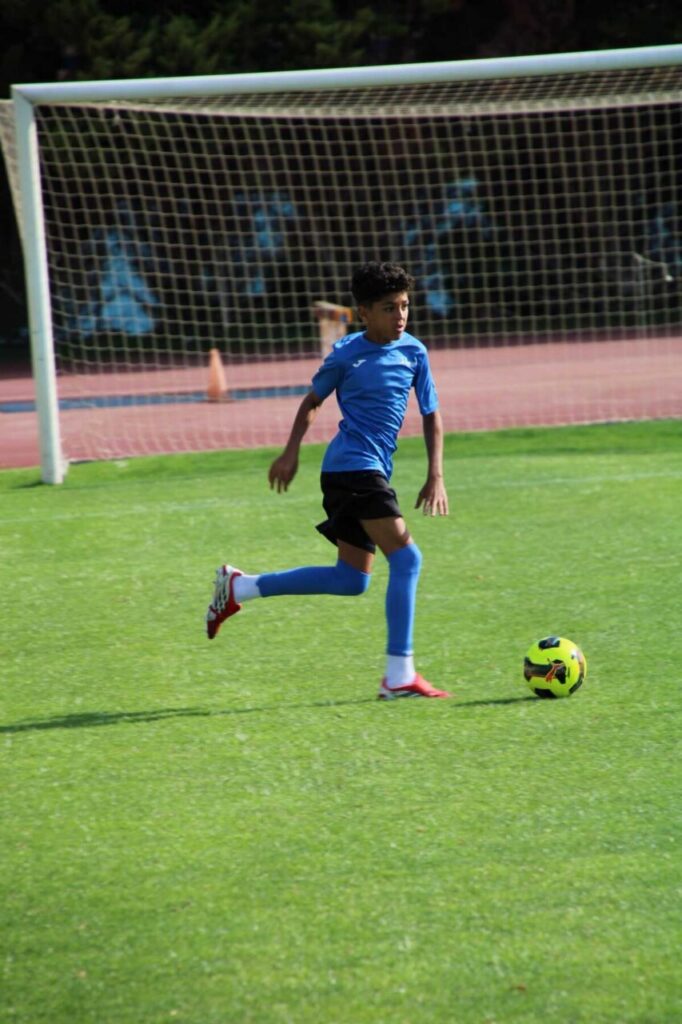 Young soccer player in a blue shirt and blue socks dribbles a yellow ball on a grassy field near the goal.