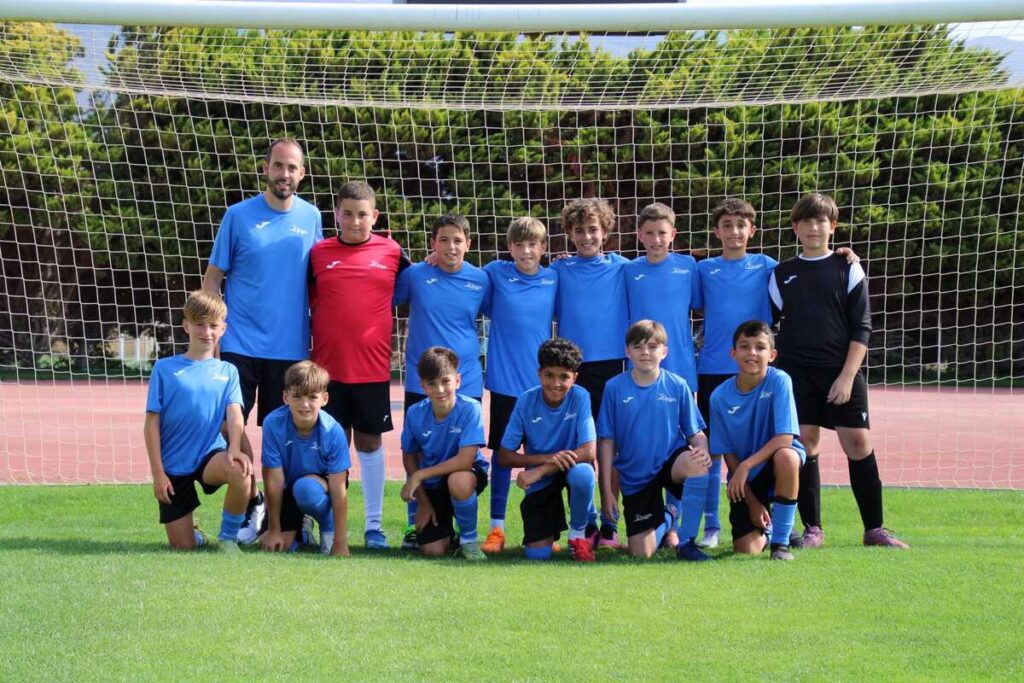 Youth soccer team photo: coach and players in blue jerseys pose in front of a goal on a sunny field, goalie in red.