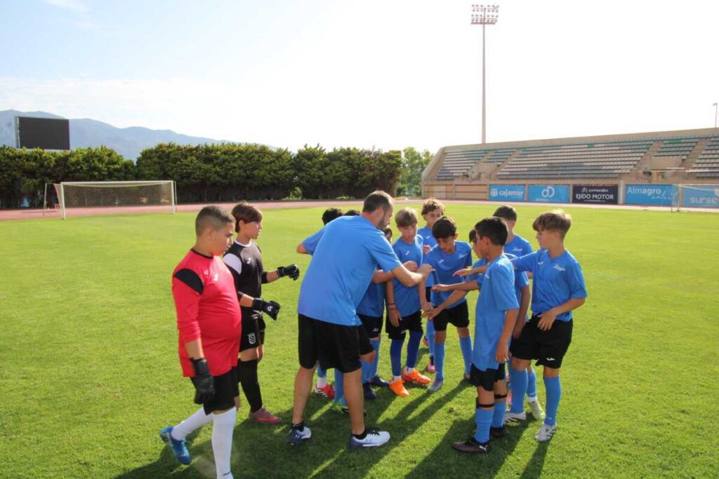 Youth soccer team in blue jerseys huddles with coach for a pre-game talk on a sunny field.
