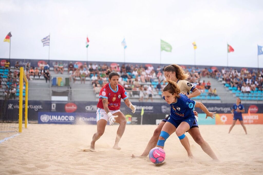 Women’s beach soccer: a blue-team player dribbles a pink ball while a defender in red pursues on sandy ground with stands in the background.