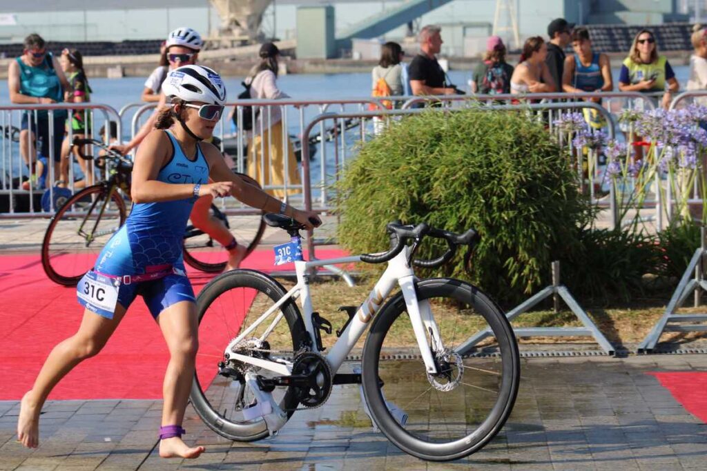 Female athlete in a blue triathlon suit and helmet runs with a white road bike beside a red carpet, spectators behind metal barriers by the water-side venue.