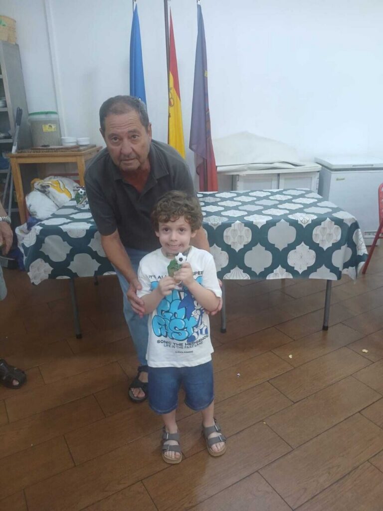 Young boy and an older man pose together in a room with flags in the background, boy holding a small trophy.