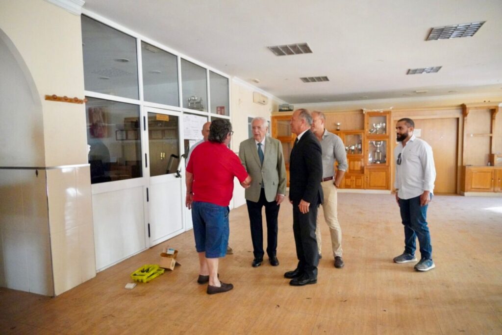 Five adults stand in a spacious room; two men in suits shake hands while others look on, with glass doors and wooden cabinets in the background.