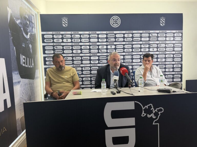 Three men sit behind a press conference table with microphones, backdrop covered in sponsor logos behind them and a large banner in the foreground.