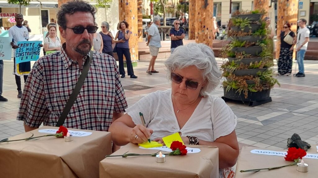 Older woman signs notes on a yellow sticky pad at a table with flowers while a sunglasses–wearing man watches in a sunny plaza; protest signs in the background.