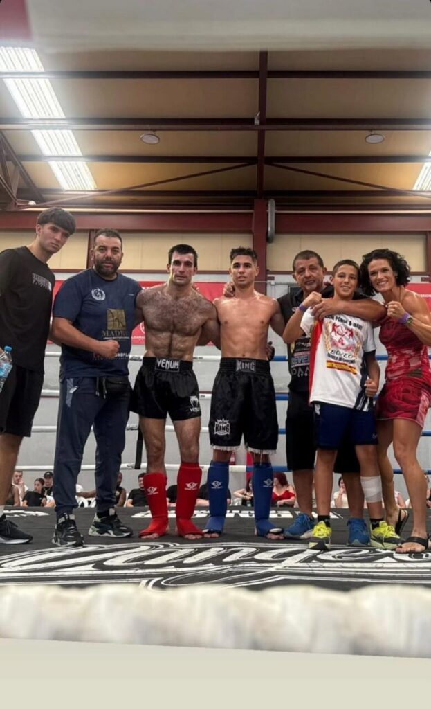 Group of boxers and coaches posing in a ring after a match, with three fighters in shorts and colored shin guards flanked by teammates and a young spectator on the right.