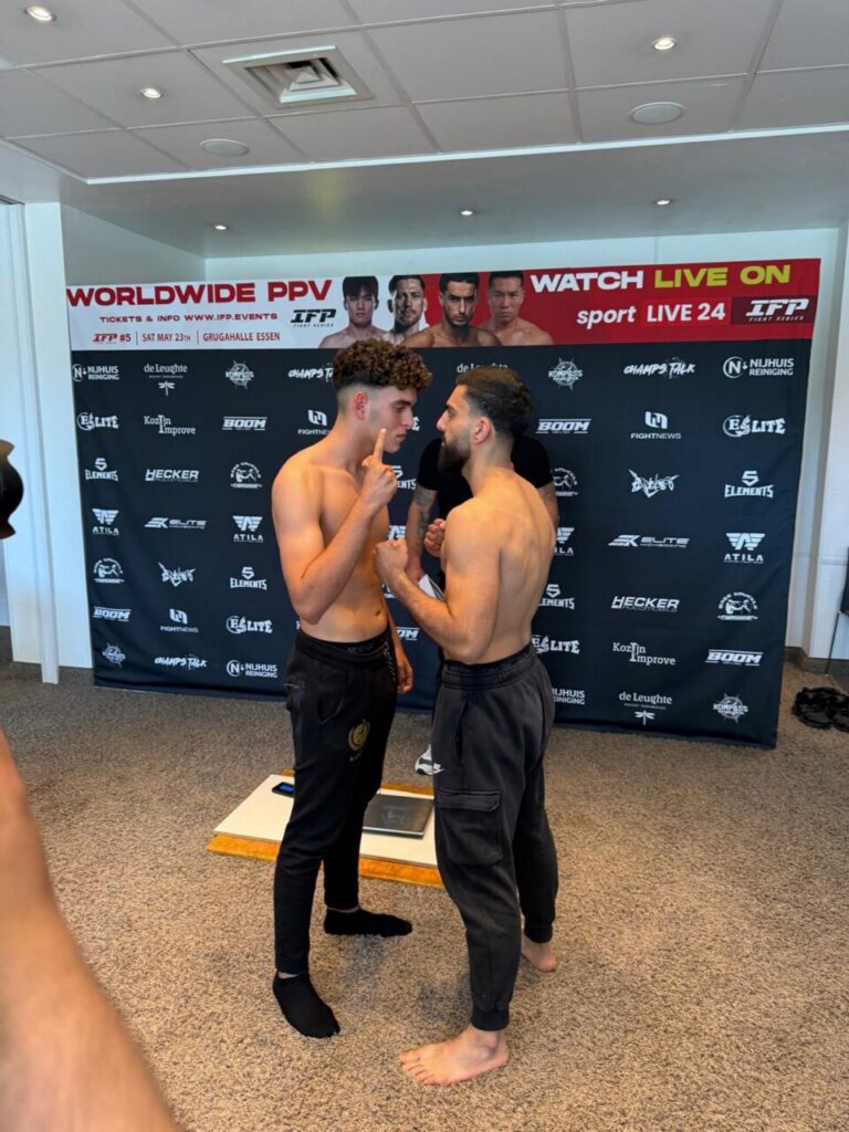Two shirtless male fighters face off in a pre-fight weigh-in, standing barefoot on a platform in front of a sponsor-filled backdrop.