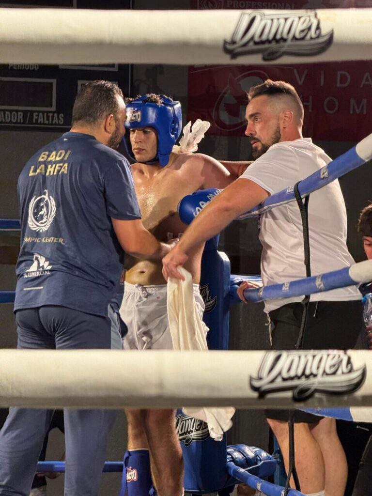 Boxer in blue headgear sits against ropes while two men attend him inside the ring, one holding a towel.