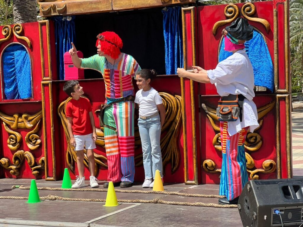 Clown in a bright striped outfit with a red wig talks to two children on a colorful red stage during a street performance.