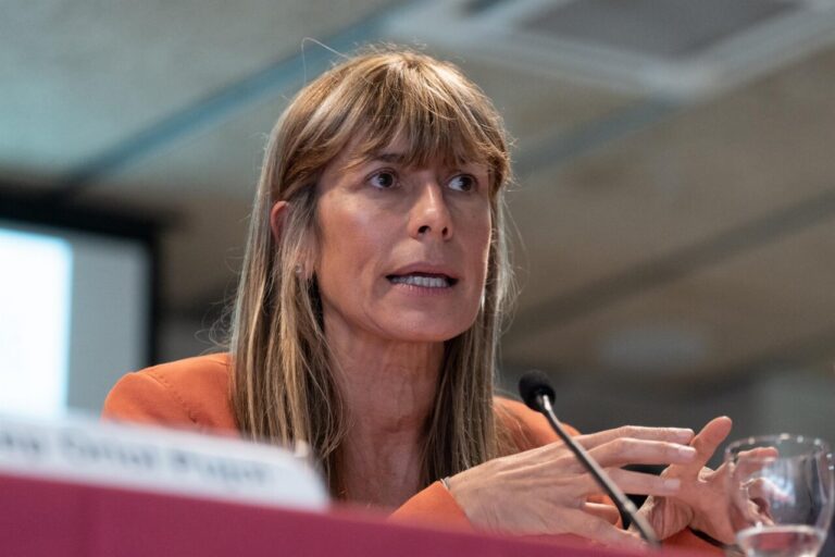 Panelist in an orange blazer speaks passionately, gesturing with her hands into a microphone on a conference table