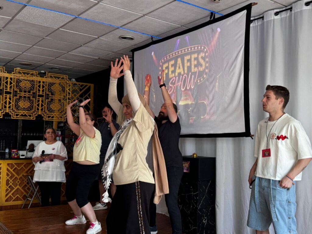 Group on stage with hands raised in a dance, FEAFES Show projected behind them.