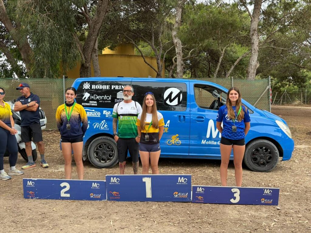 Three young female athletes stand on a podium with medals; 1st, 2nd, and 3rd place boards in front, blue sponsor van behind them.
