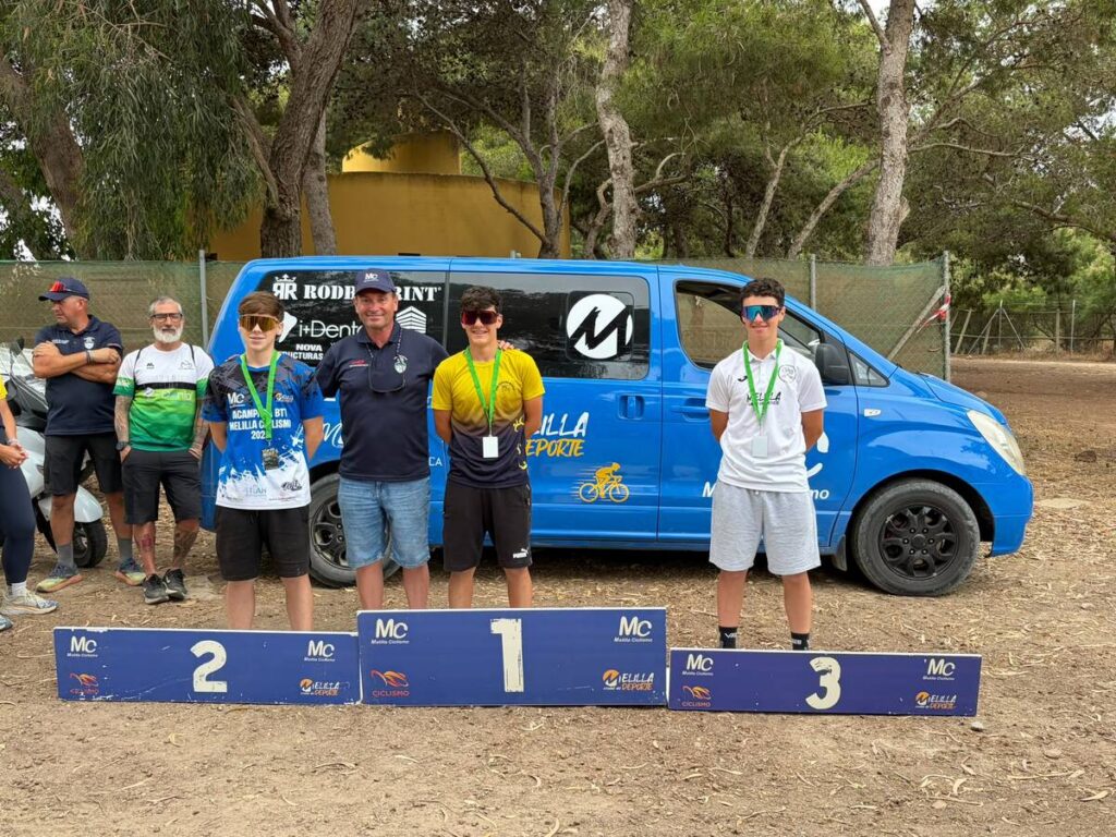 Three young cyclists stand on a podium with numbered placards (2, 1, 3) and medals, in front of a blue van on a dirt lot.