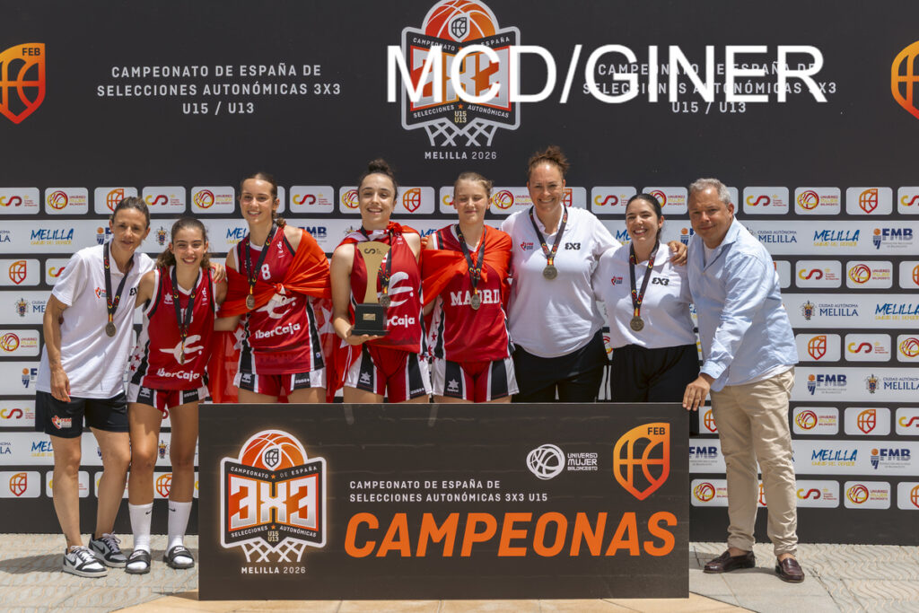 Women’s 3x3 basketball team posing with medals and trophy on stage, red uniforms, coaches beside them, banner reads CAMPEONAS and 3x3 Melilla 2026
