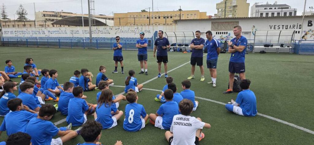 Group of young soccer players in blue uniforms sit on a turf field listening to six coaches standing in a line in front of them on the sideline.