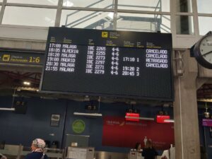 Large departure board at a station showing times and destinations (e.g., Malaga, Madrid) with some statuses like CANCELADO and RETRASADO/OBSERVACIONES nearby.