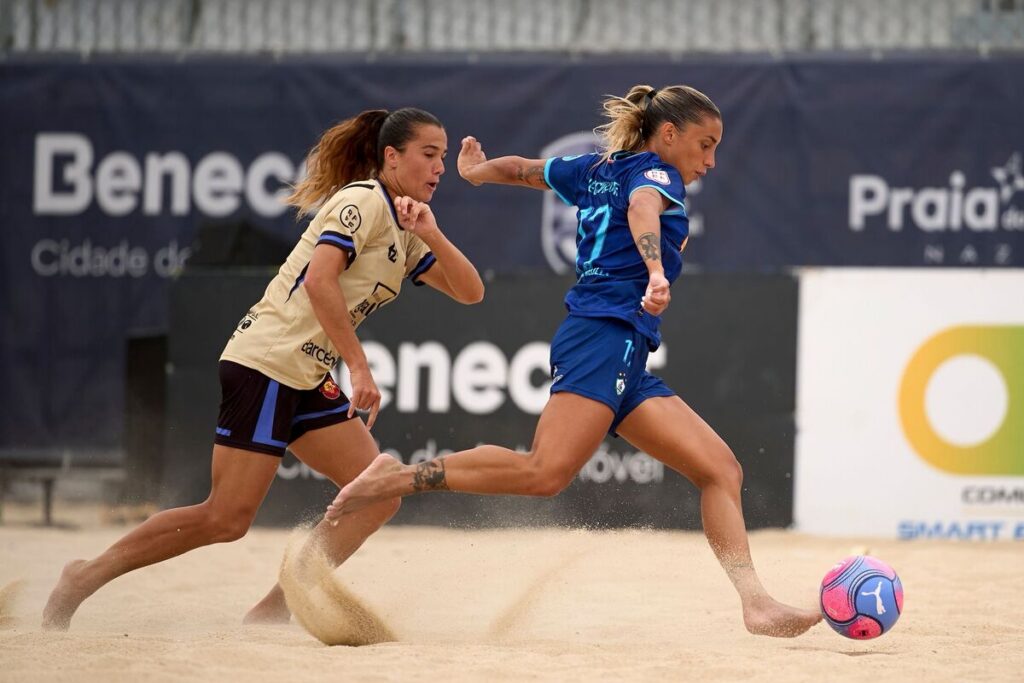 Two female beach soccer players chase a pink-and-blue ball on a sandy pitch, one in beige jersey and the other in blue, sprinting side by side.