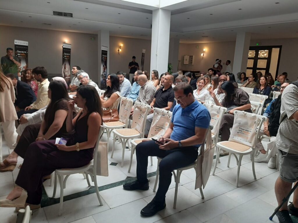 Audience seated in a bright conference room, facing forward as a presentation takes place in the front area, some participants looking at devices.